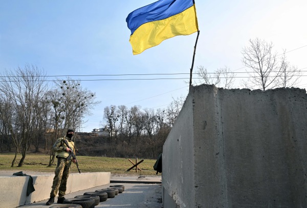 图为2022年3月28日，乌克兰首都基辅郊区的一个检查站，一名乌克兰士兵在持枪把守，检查站上飘扬着一面乌克兰的蓝黄两色的国旗。（图片来源：SERGEI SUPINSKY/AFP via Getty Images）