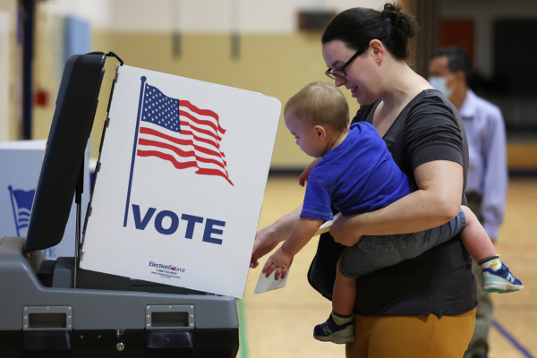 图为2022 年 6 月 21 日，美国弗吉尼亚州亚历山大（Alexandria）市举行的中期选举初选日里，一名选民抱着她的小孩在一个投票站里投票。弗吉尼亚州在从摇摆州转向越来越倾向共和党。（图片来源：Alex Wong/Getty Images）