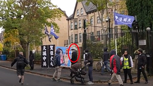 10月16日，居英港人在英国曼城中领馆外抗议，一中年人（红圈）从中领馆内走出，踢倒示威直幡，抢走习近平漫画海报。（图片来源：视频截图）