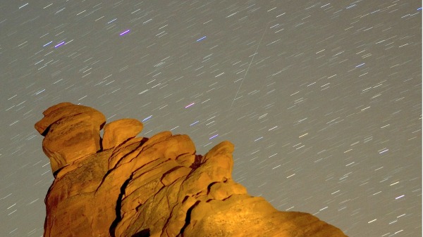 2007年12月14日，在美国内华达州火谷州立公园，一颗双子座流星斜划过天空，映衬着七姊妹岩层之一的山峰上空的星迹。这次流星雨被称为双子座流星雨，因为它似乎是从双子座的北河二恒星附近辐射出来的，被认为是由一颗名为 3200 Phaethon 的类小行星天体抛射出的碎片造成的。每年十二月都会出现阵雨。 （Ethan Miller/Getty Images)